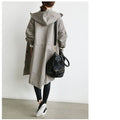 Person wearing a long gray coat with a hood, holding a black bag against a white background