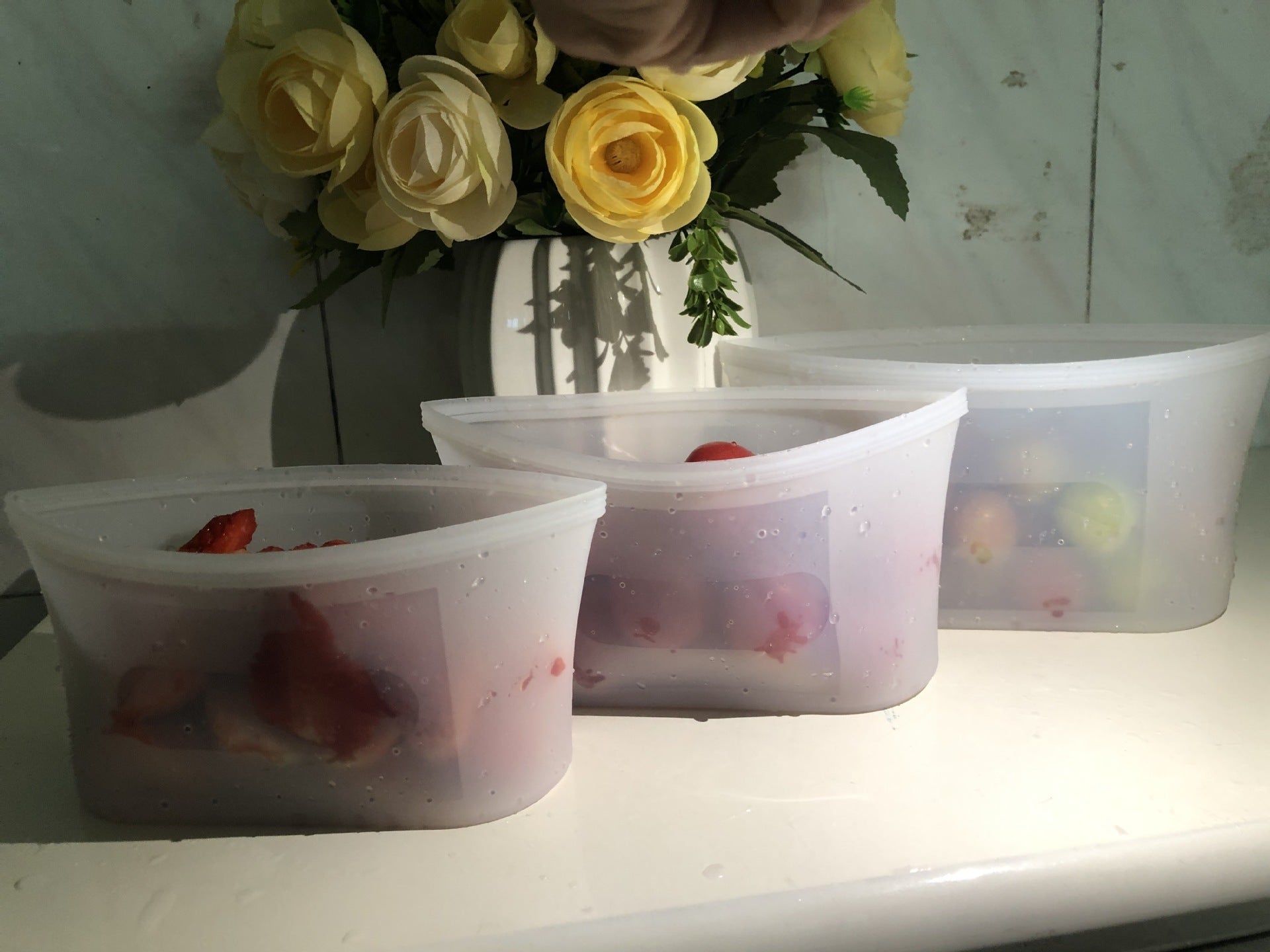 Three silicone food wraps with strawberries on a surface, with a vase of yellow flowers in the background; Reusable silicone food storage bags zip seal leakproof