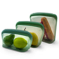 Set of three green silicone food storage containers with fruits and a sandwich inside on a white background; Reusable kitchen silicone bags for meal prep and leftovers