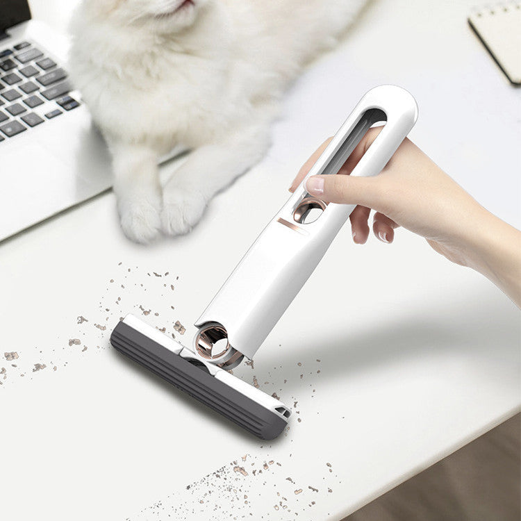 Hand using a small handheld vacuum cleaner on a desk with a cat in the background