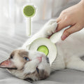 Cat being petted with a green and white pet grooming brush on a soft surface.