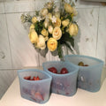 Three blue silicone food storage containers with fruits on a white surface, with a bouquet of yellow roses in the background